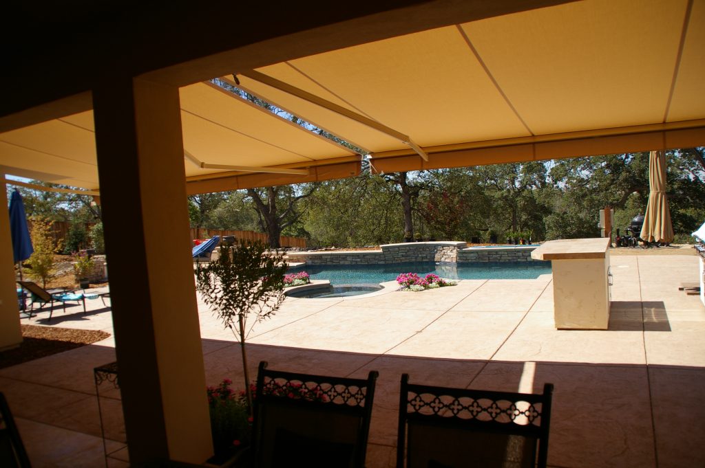 View of sunlit backyard pool from beneath a goldenrod retractable awning.
